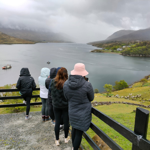 Students standing with coats looking out a body of water. Grey clouds circle in the sky.